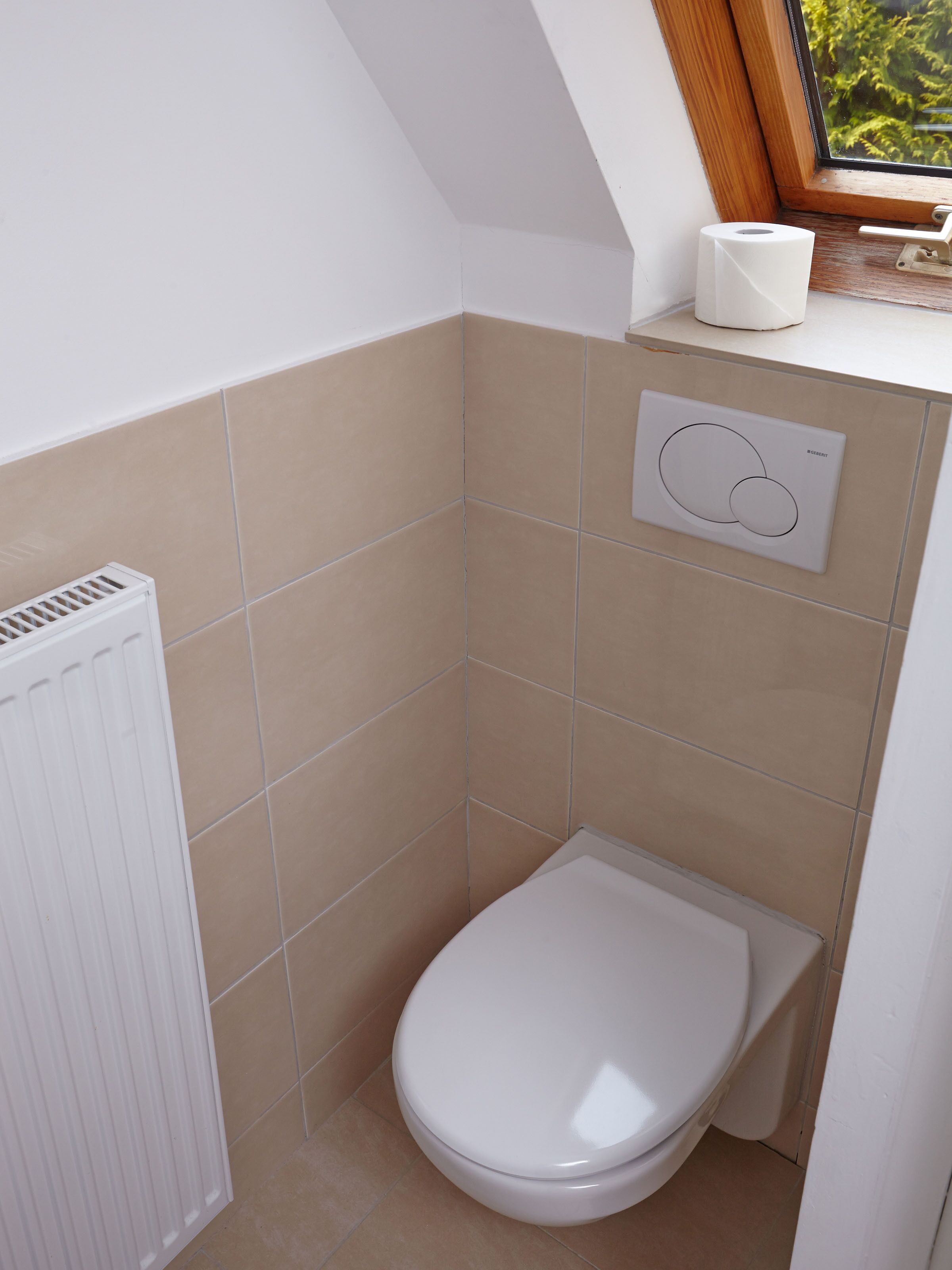 Une petite salle de bain moderne, équipée de toilettes, d’un radiateur et d’une fenêtre donnant sur un espace verdoyant.