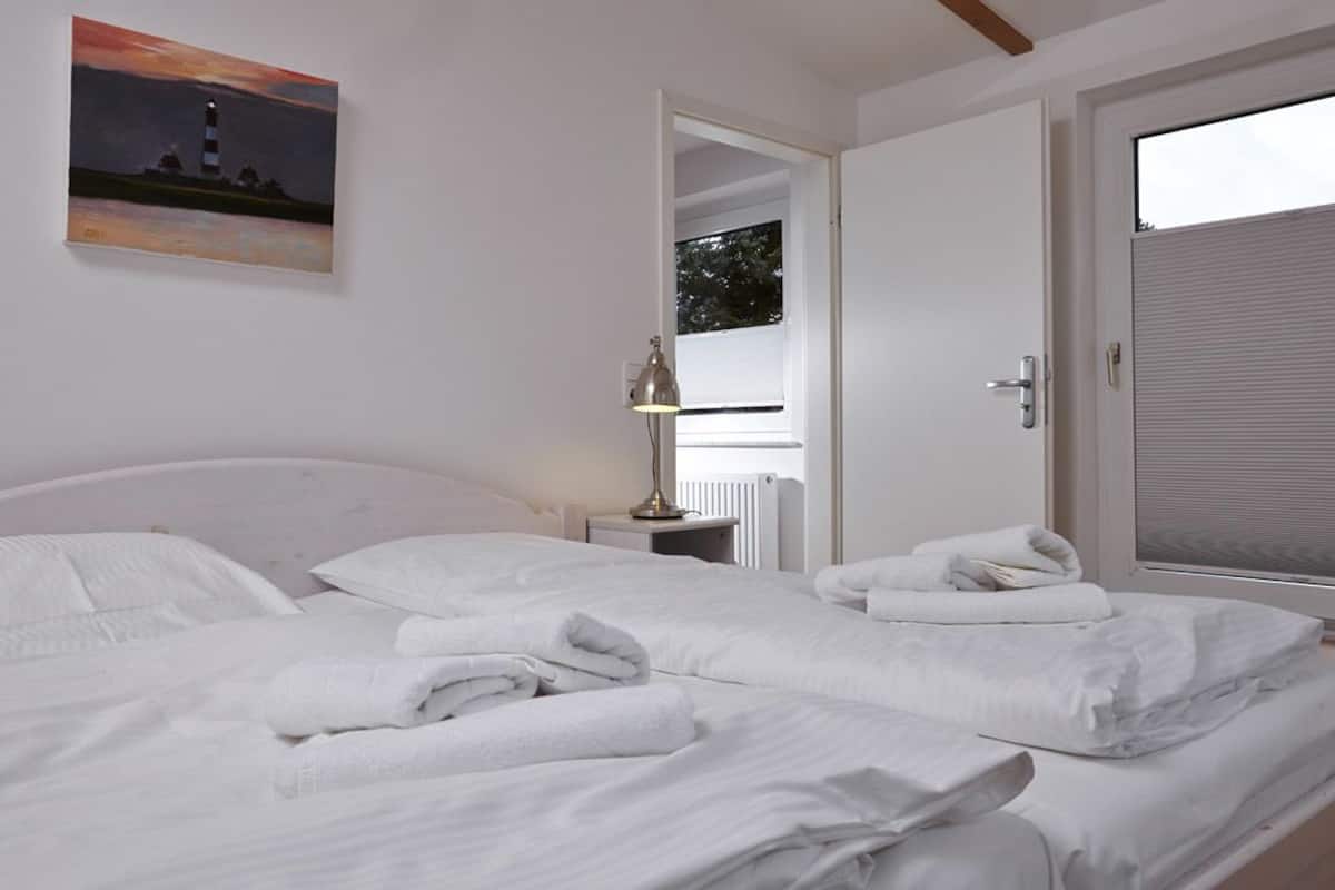 Ein Hotelzimmer mit zwei Betten, weißen Leinentüchern, einem Gemälde eines Leuchtturms, einer Lampe und Blick durch eine Tür.