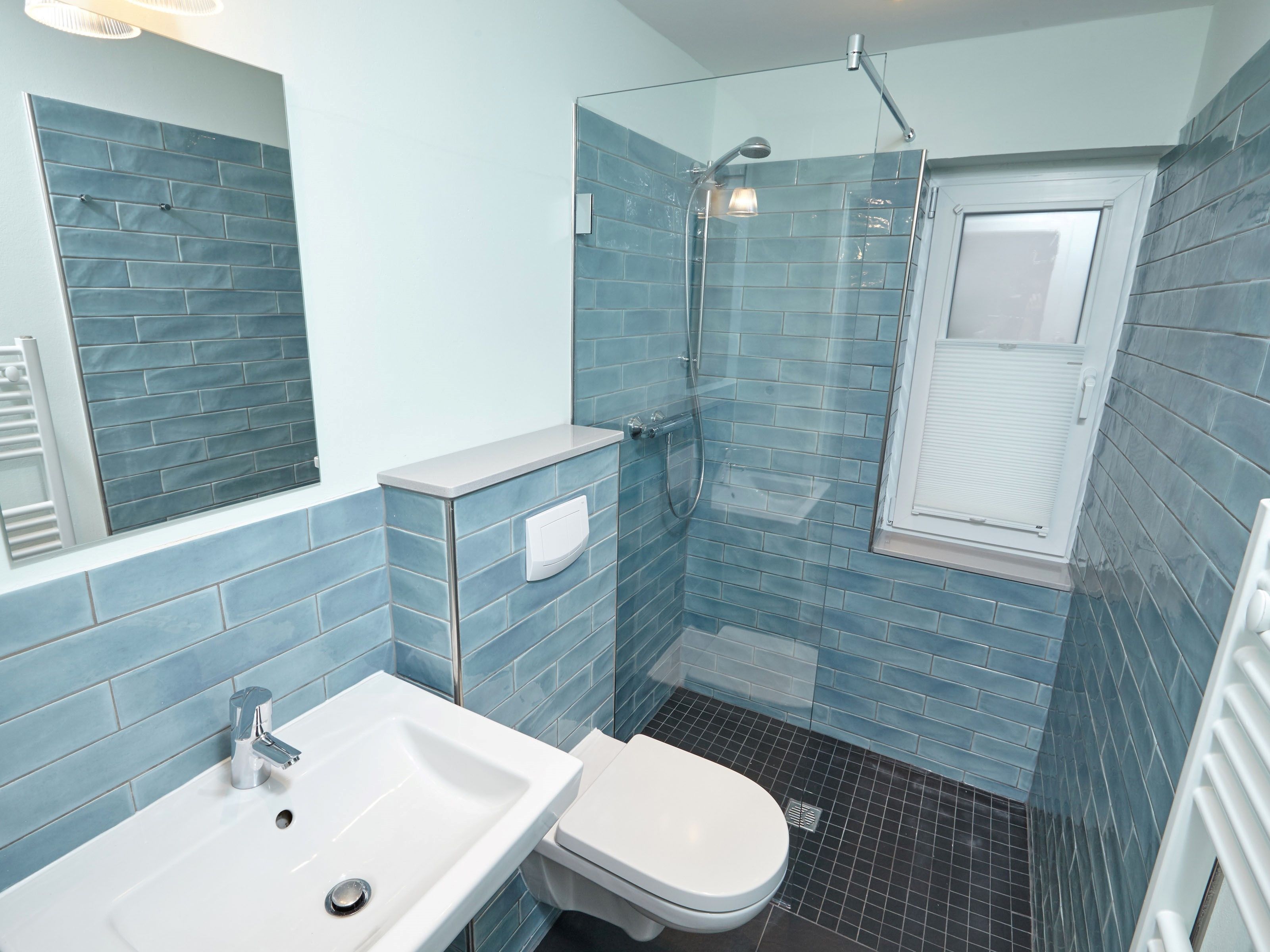 A bathroom with a sink, toilet, and shower area, featuring blue-tiled walls and a window.