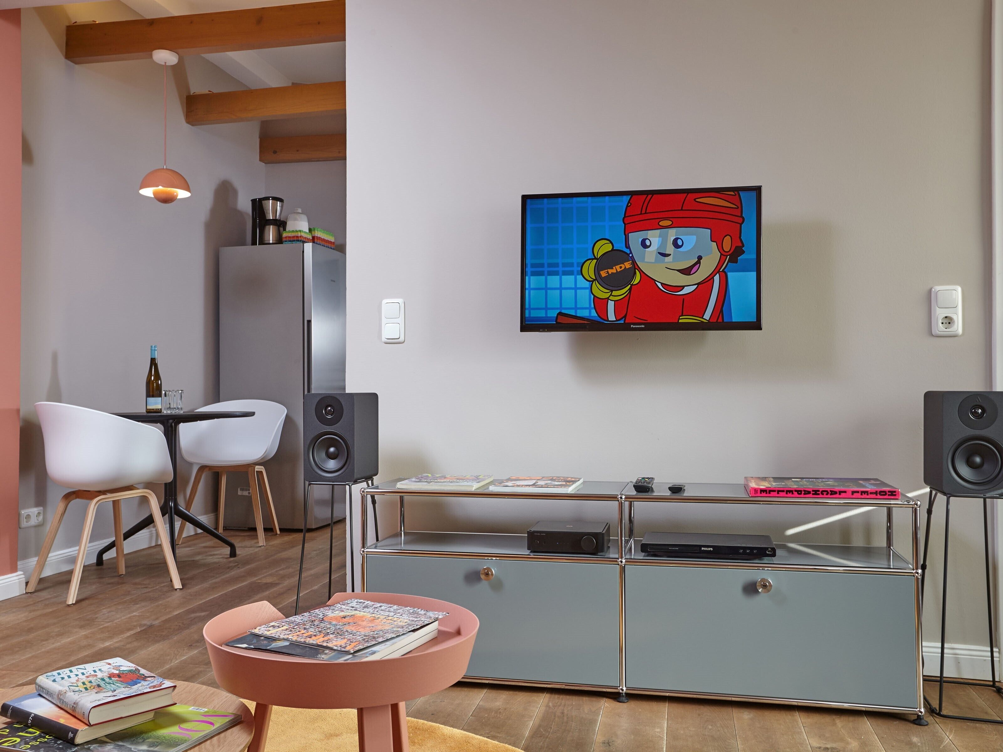 Ein modernes Wohnzimmer mit Flachbildfernseher, einer Kaffeemaschine, einem Bücherregal und einem Essbereich mit Stühlen.