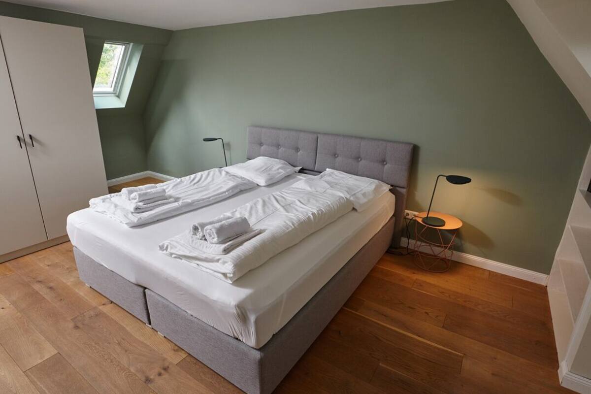 Ein ordentlich bezogenes Bett mit weißen Leinen und Handtüchern, ein grauer Kopfteil, ein kleiner Tisch mit einer Lampe und ein Kleiderschrank.