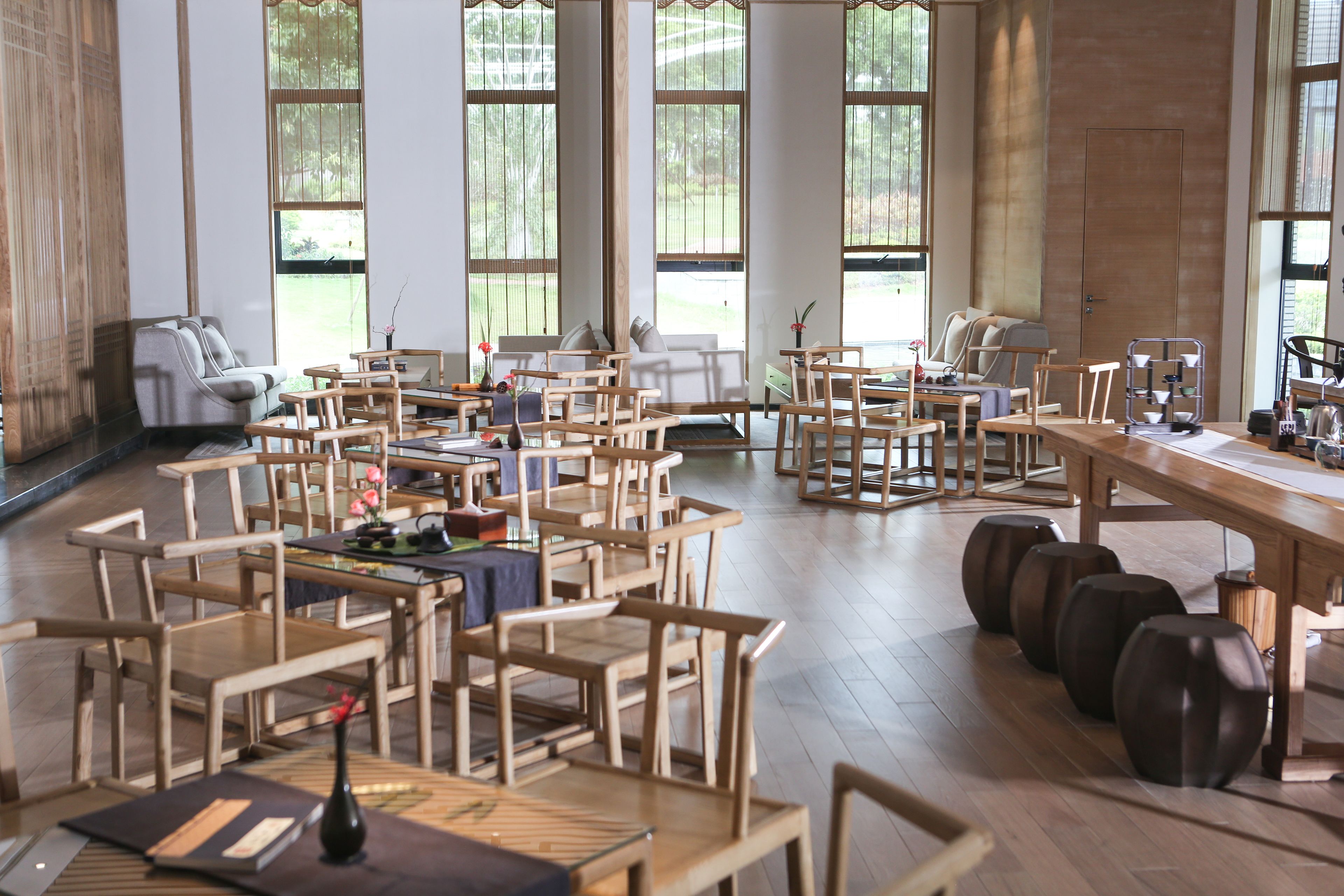 A spacious room with multiple wooden tables and chairs arranged for dining. Large windows allow natural light to fill the space.
