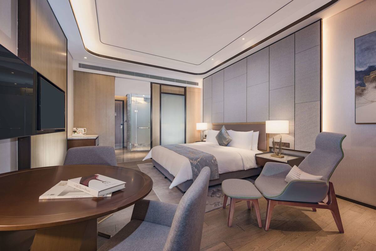 Une chambre d’hôtel moderne dotée d’un grand lit, d’un coin salon avec une table et des chaises, d’une télévision et d’une salle de bain visible à travers une porte en verre.