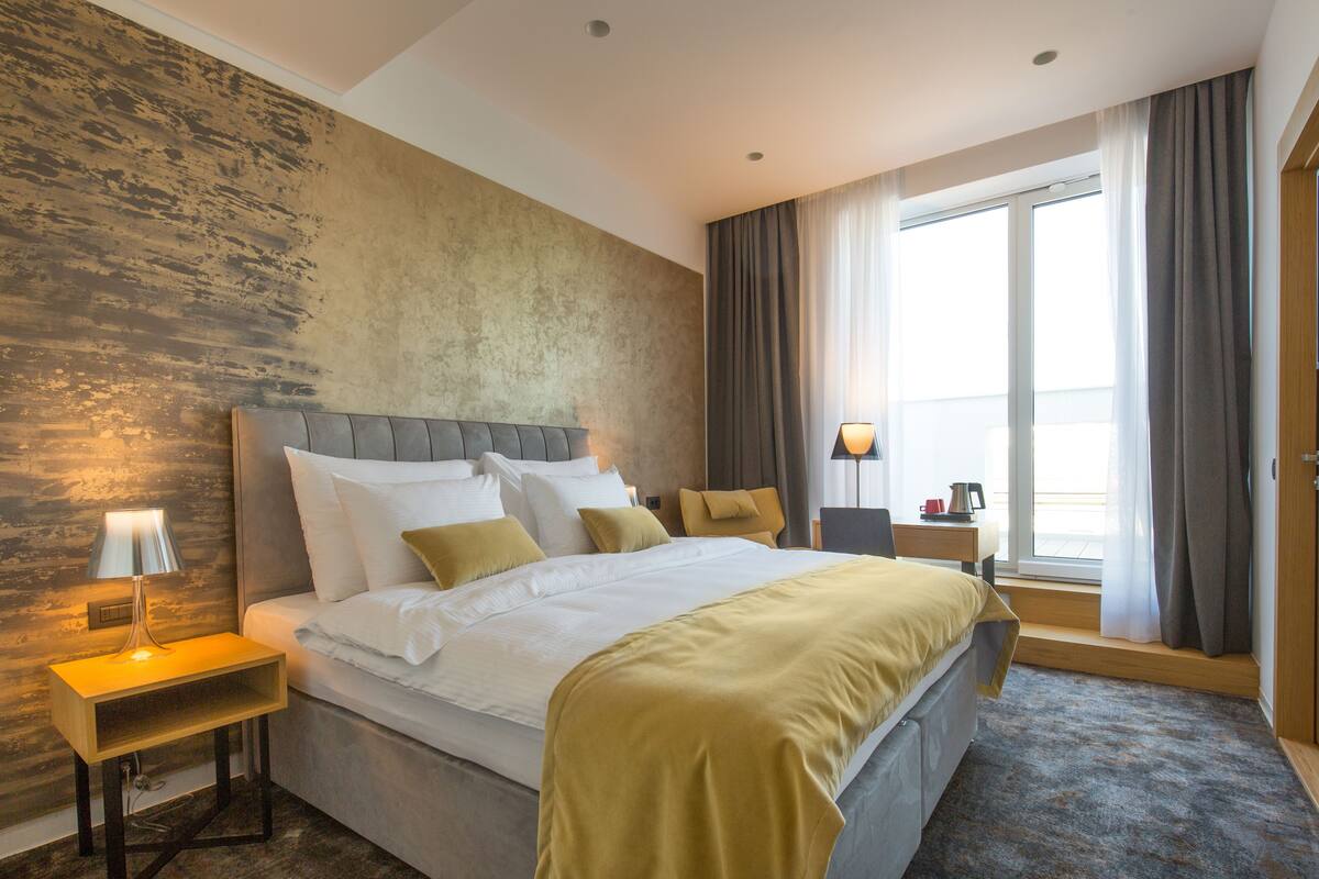 Una habitación de hotel moderna con una cama grande, una mesita de noche con lámpara, una ventana con cortinas y una pared de madera.