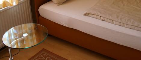 A small, single-bed room with a glass table, a radiator, and a painting on the wall.