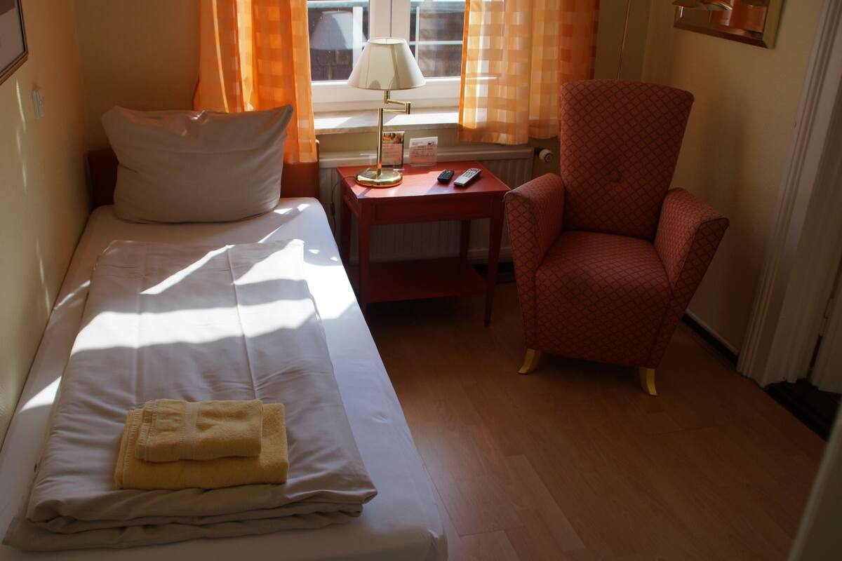 Une petite chambre avec un lit simple, un bureau et une chaise, une fenêtre avec des rideaux et un lit avec une serviette pliée.