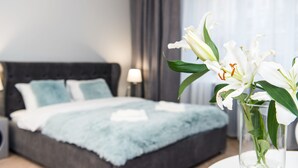 Een moderne slaapkamer met een donker hoofdbord, een zachte blauwe deken en een vaas met witte lelies op een nachtkastje.