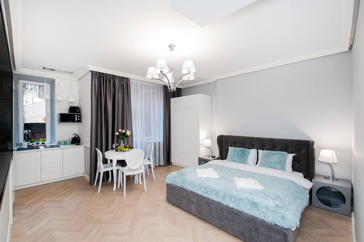 A modern hotel room with a large bed, a dining area with a table and chairs, a kitchenette, and a separate bathroom.