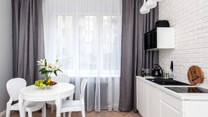 Een moderne keuken met witte kasten, een ronde eettafel met stoelen, een schaal fruit en een raam met dunne gordijnen.