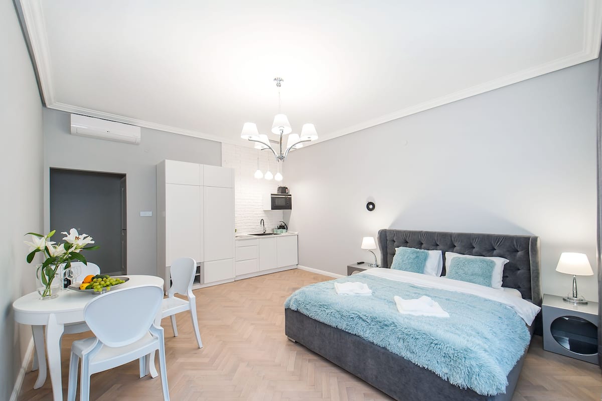 A modern bedroom with a large bed, a dining area with a table and chairs, and a kitchenette in the background.