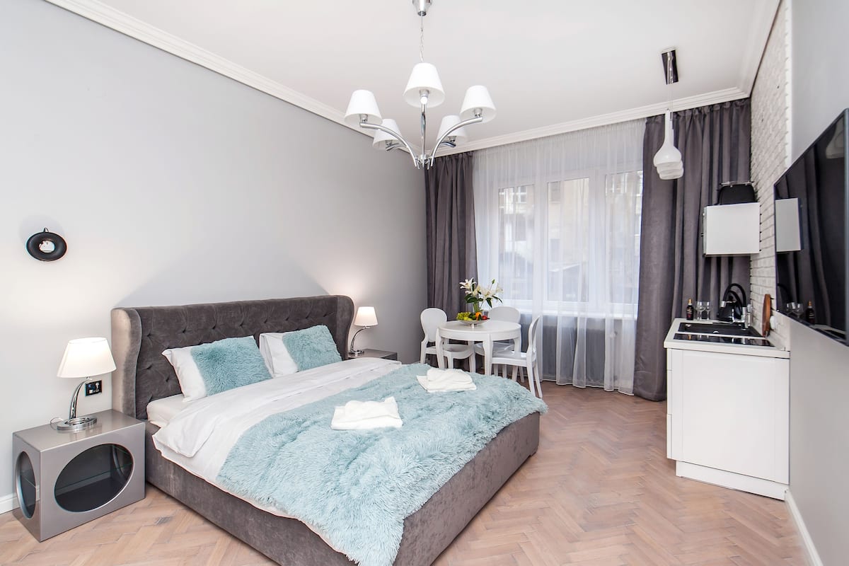 A modern bedroom with a large bed, bedside tables, a small kitchen area, and a dining table with chairs.