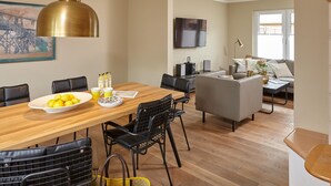 Uma sala de estar moderna com uma mesa de jantar de madeira, cadeiras pretas, um sofá cinzento e uma televisão montada na parede.