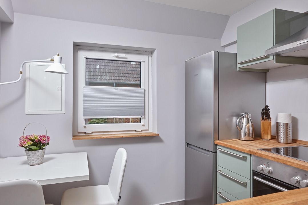 Une cuisine moderne dotée d’un réfrigérateur encastré, d’un plan de travail en bois, d’une fenêtre avec des stores et d’une chaise blanche.