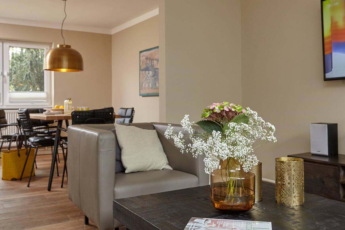 Ein modernes Wohnzimmer mit einem grauen Sofa, einem Couchtisch mit Blumenvase, einem Essbereich mit Tisch und Stühlen sowie einem großen Fenster.