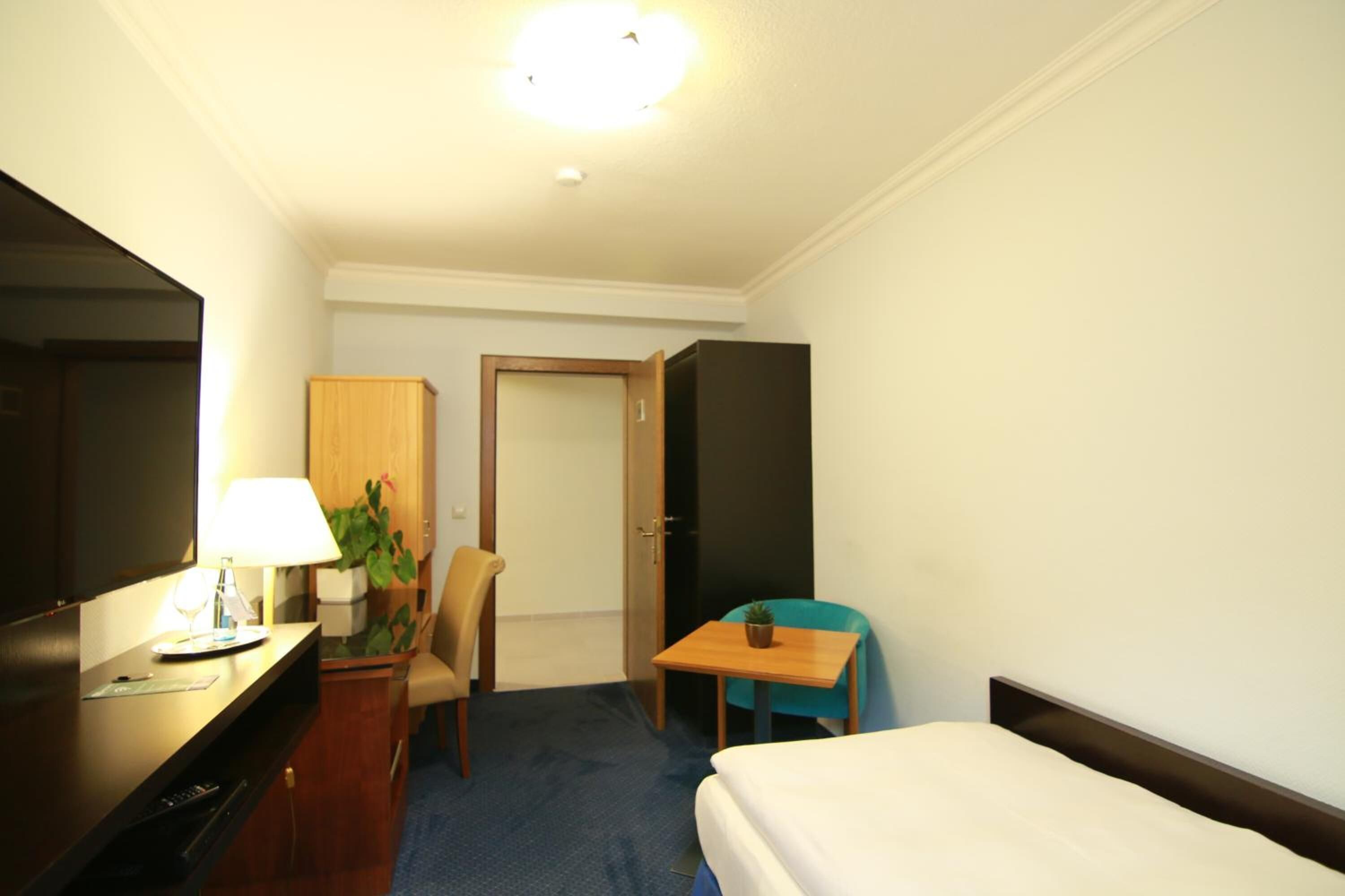 A hotel room with a bed, a desk with a lamp, a chair, a small table with a plant, and a wardrobe.