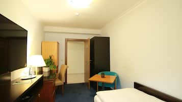 A hotel room with a bed, a desk with a lamp, a chair, a small table with a plant, and a wardrobe.