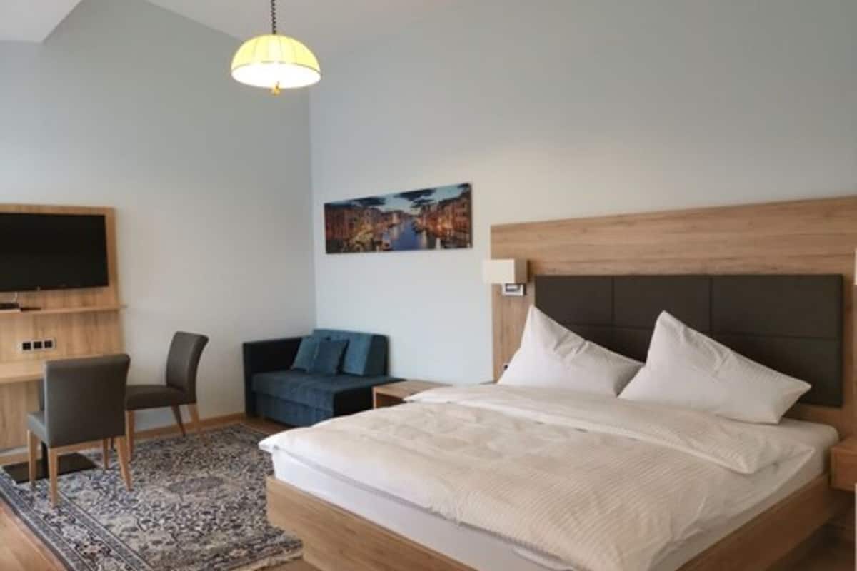 A modern hotel room with a large bed, a desk, a chair, a television, and a painting on the wall.