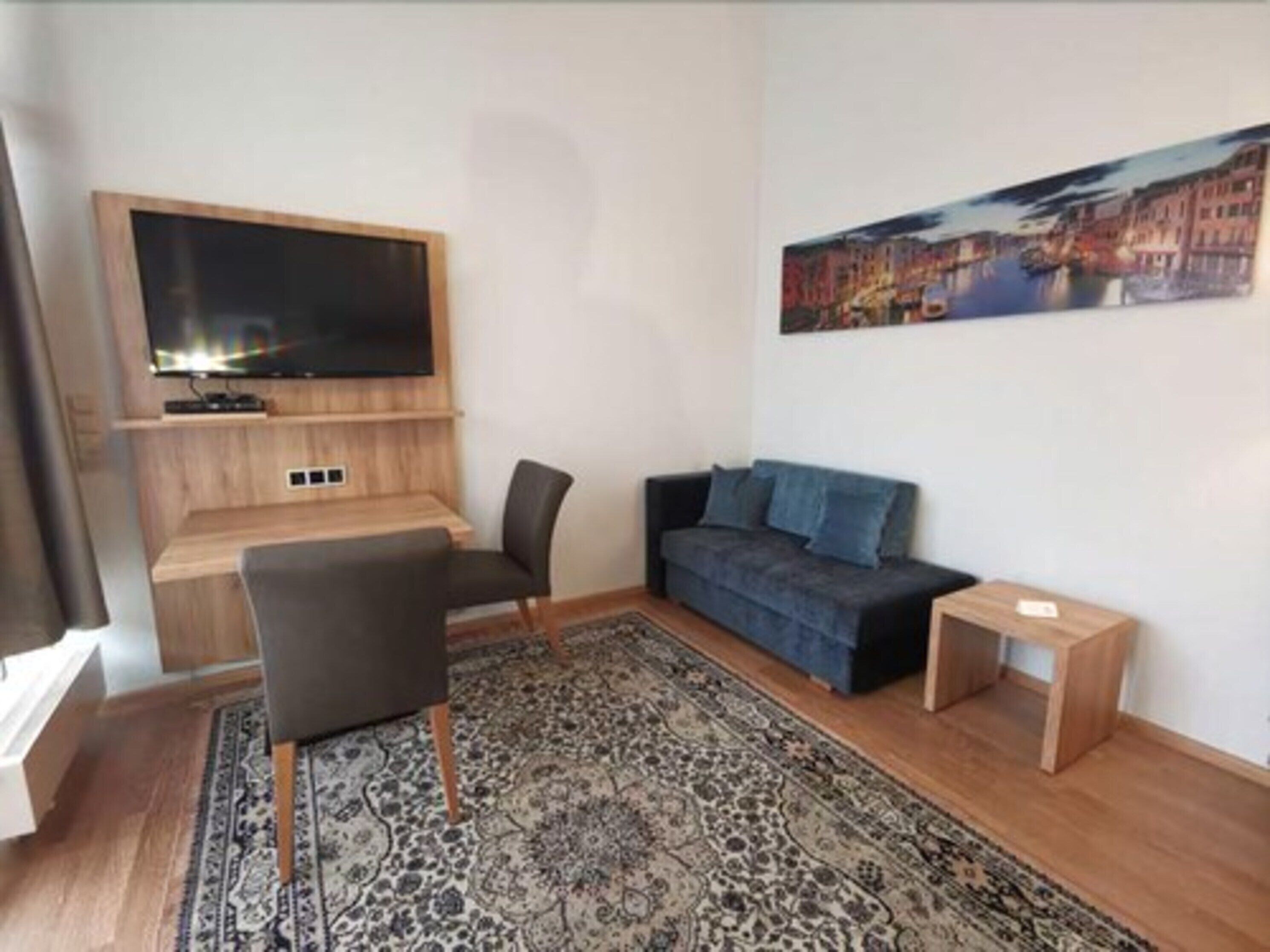 Una sala de estar moderna con un televisor de pantalla plana montado en una pared de madera, un sofá azul oscuro, una alfombra estampada y una pequeña mesa auxiliar de madera.
