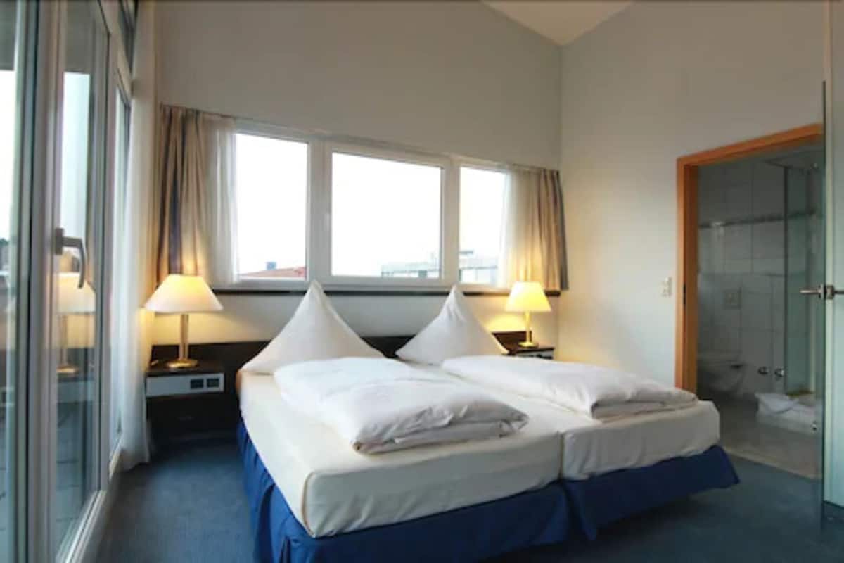 A hotel room with a double bed, two bedside tables with lamps, a large window with curtains, and a glass shower enclosure.