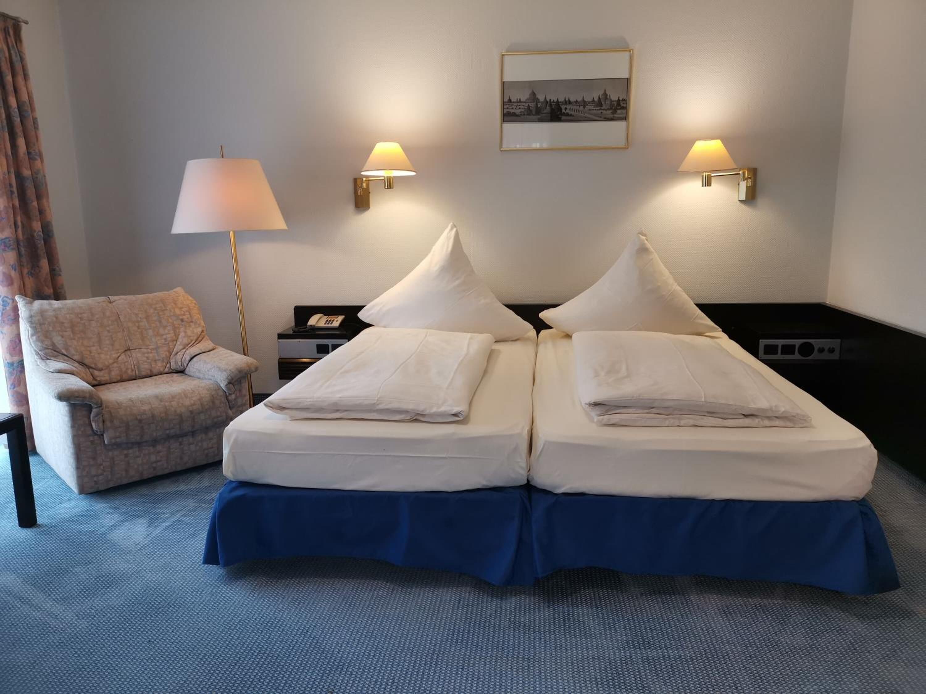 Quarto de hotel com duas camas, uma cadeira, escrivaninha com abajur e um quadro na parede.