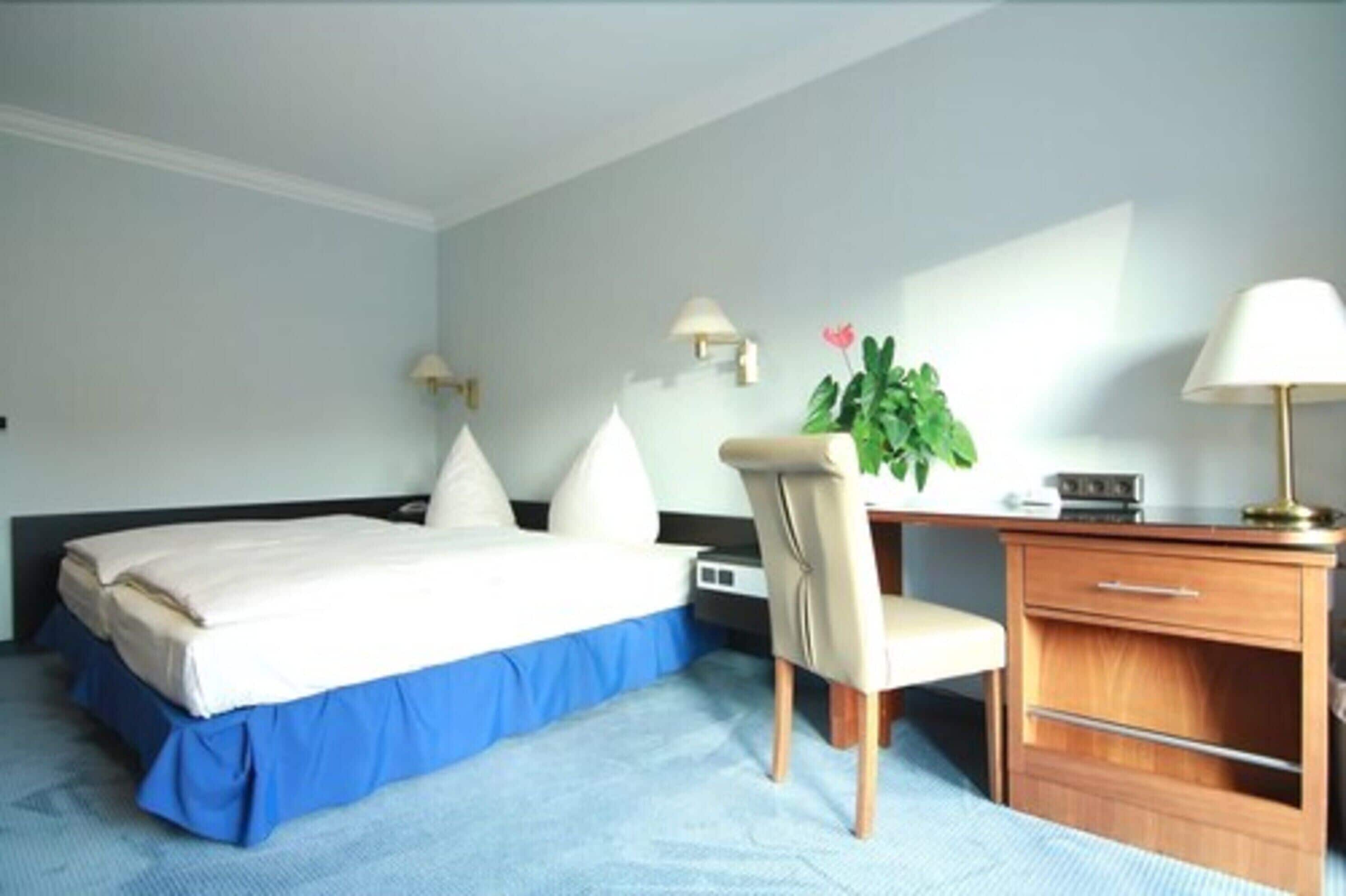 A hotel room with a bed, a desk, a chair, a lamp, a plant, and a nightstand.