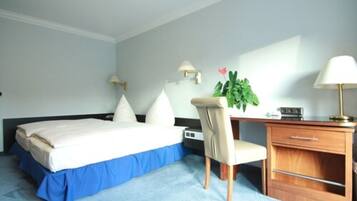 A hotel room with a bed, a desk, a chair, a lamp, a plant, and a nightstand.