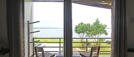 A balcony with two wooden chairs and a small table, overlooking a body of water and a grassy area with trees.