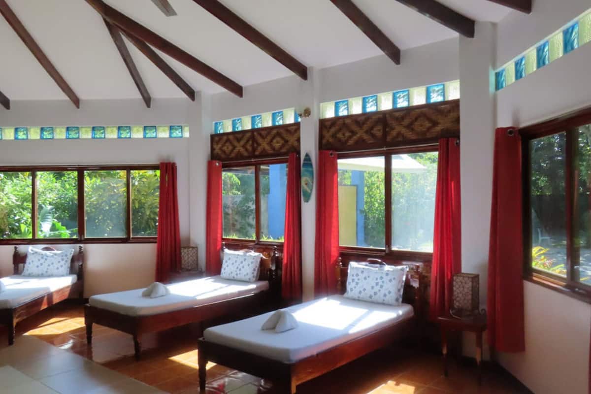 Una habitación con tres camas de madera, ventanas amplias con cortinas rojas y piso de baldosas.
