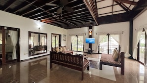 A spacious room with a large sofa, a coffee table, and a television. The room has a high ceiling with wooden beams and large windows with curtains. - Ocean 101 Beach Resort (General Luna)