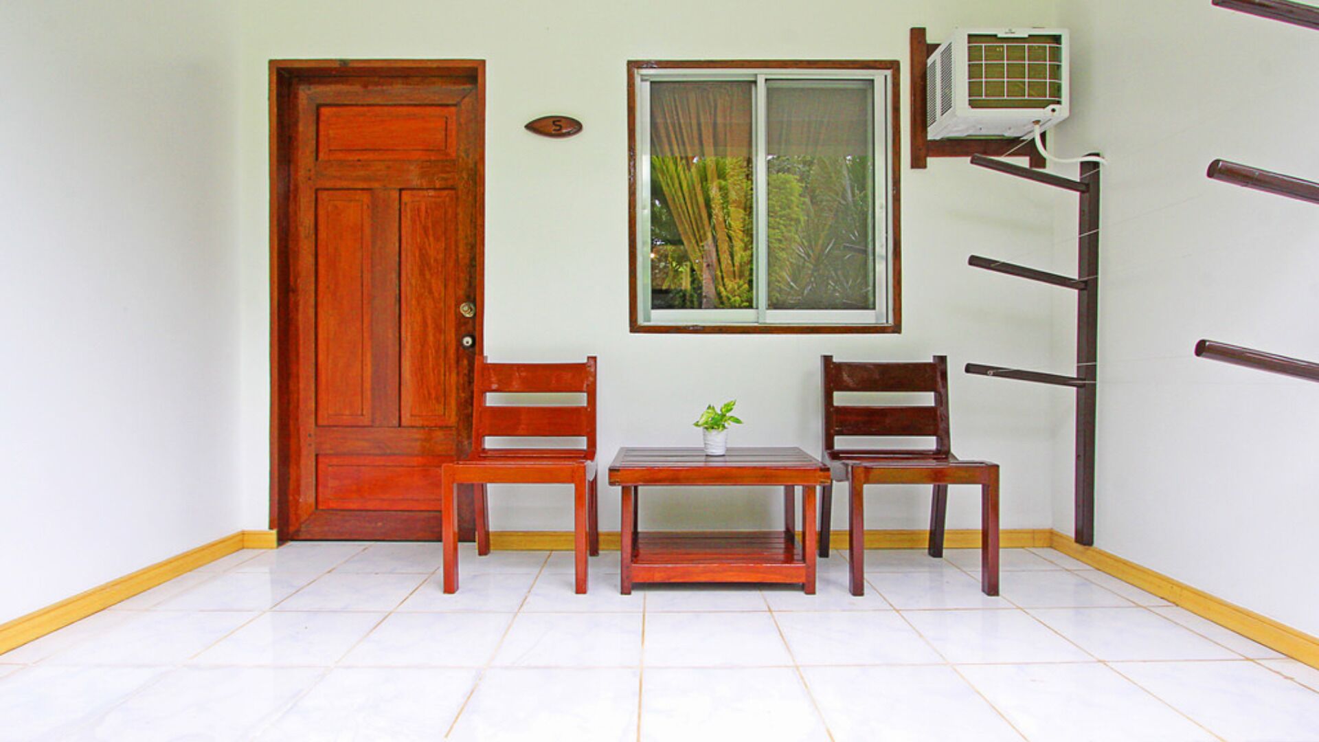 A small room with a wooden door, a window with a view of greenery, a table with a plant, and two chairs.