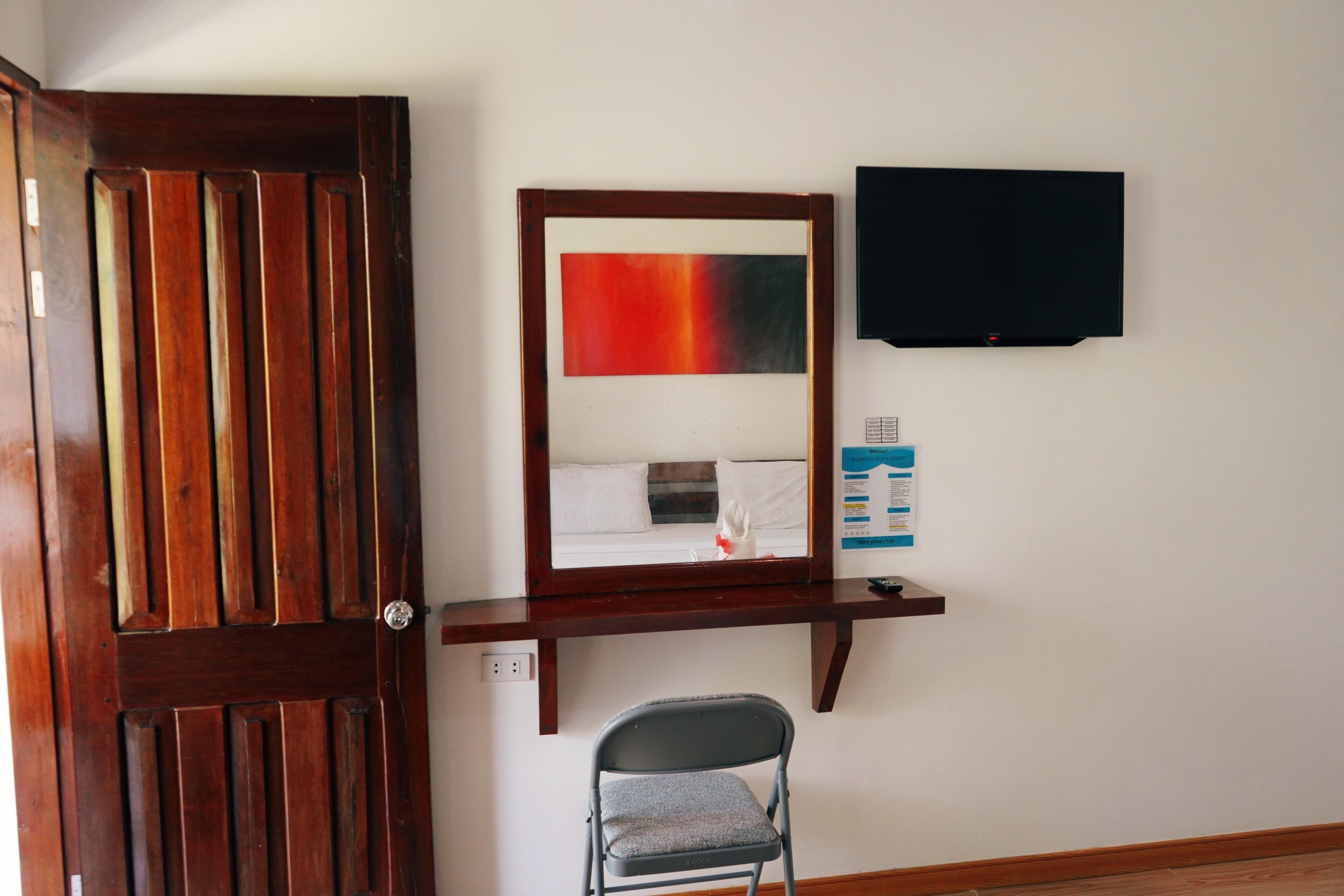 A room with a wooden door, a mirror with a red and black gradient, a flat-screen TV, a small shelf, and a folding chair.