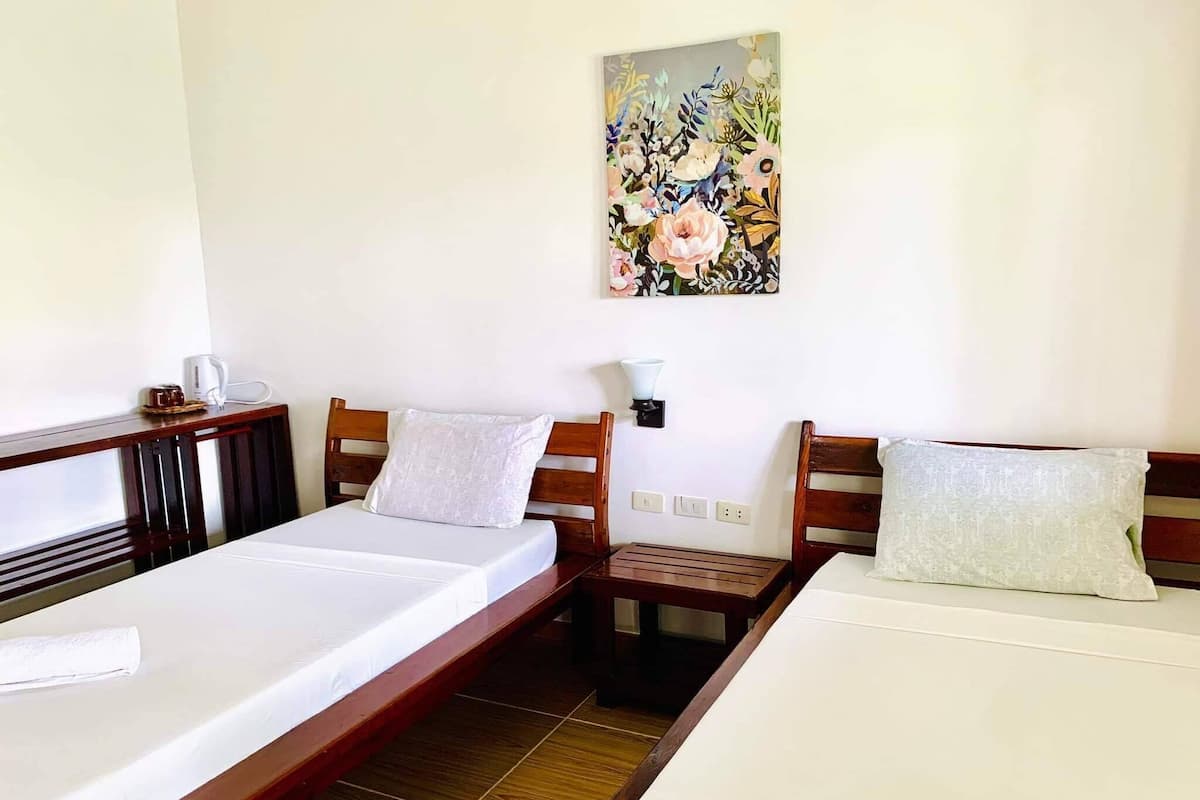 Habitación de hotel con dos camas individuales, cabecera de madera, mesita de noche, cafetera, lámpara de pared y un cuadro con motivos florales.