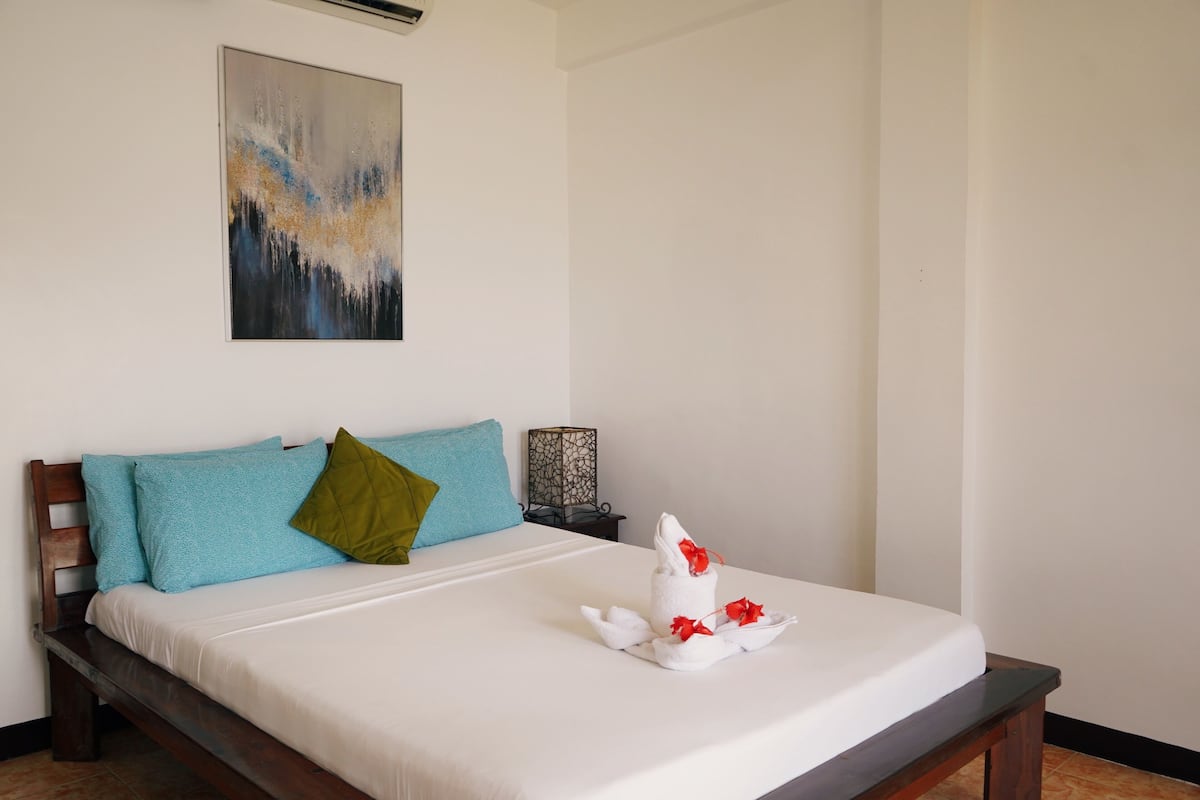 Una cama bien hecha con sábana blanca, una toalla de flores rojas, dos almohadas azules y una almohada verde.