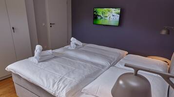 A neatly made bed with white linens and a grey headboard, a flat-screen TV mounted on the wall, and a bedside table with a lamp.
