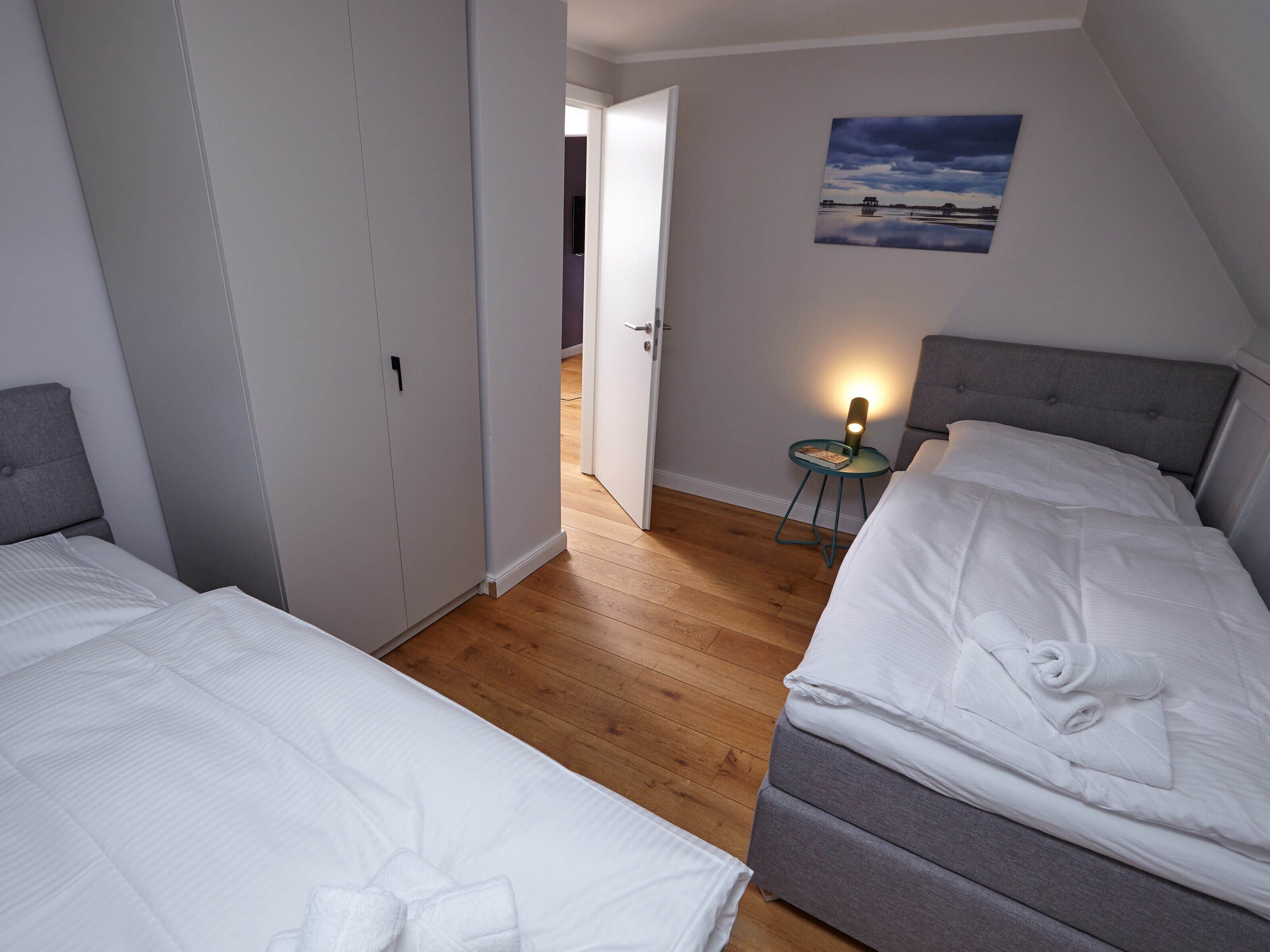 A bedroom with two beds, a small bedside table with a lamp, a painting on the wall, and a door leading to another room.