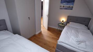 A bedroom with two beds, a small bedside table with a lamp, a painting on the wall, and a door leading to another room.