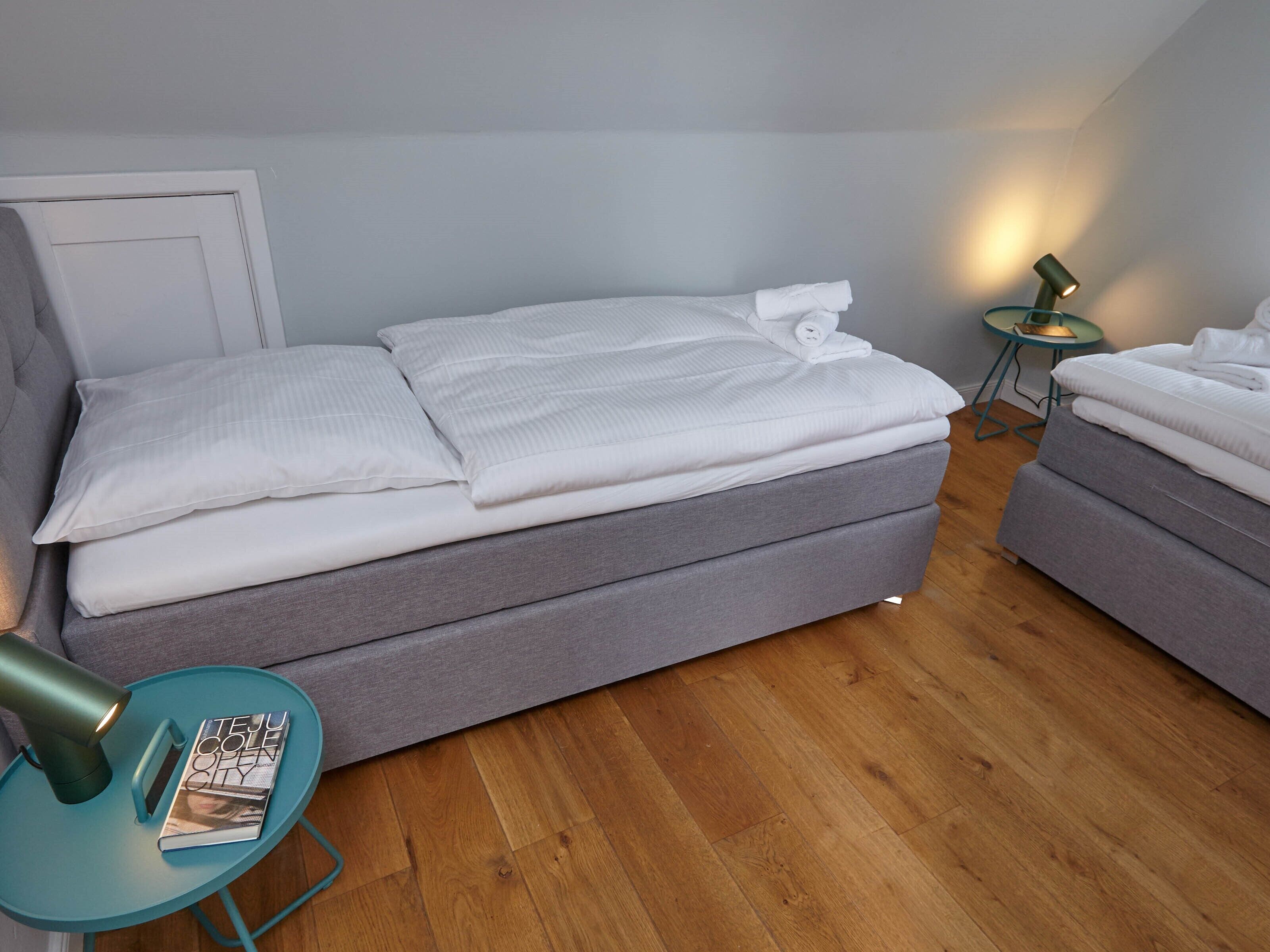 A modern bedroom with a grey sofa bed, a small teal side table with a magazine, and a lamp on a matching side table.