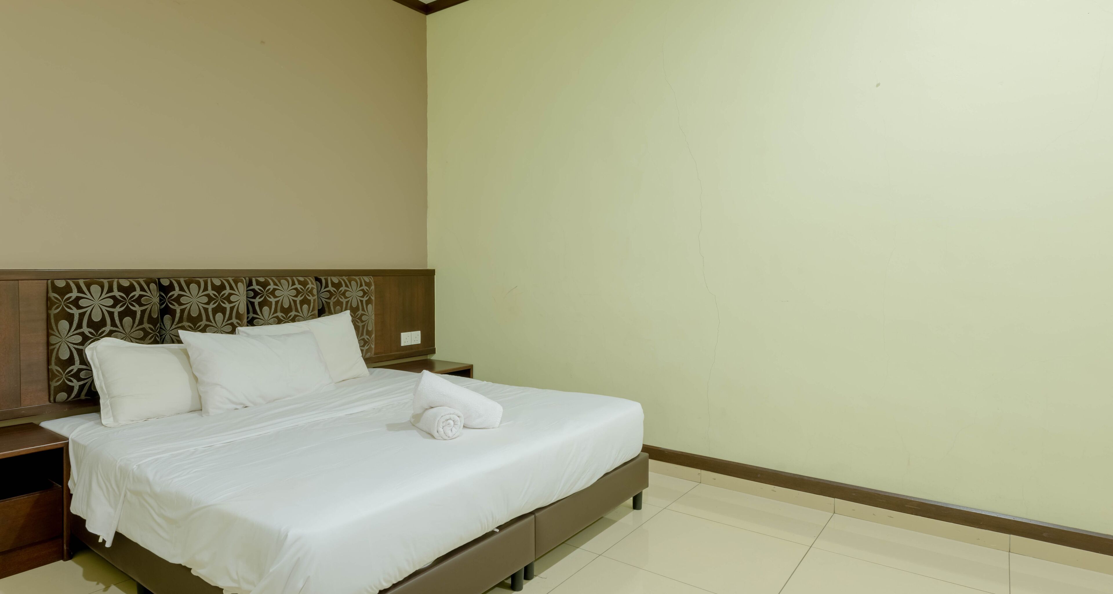Una cama bien hecha con sábanas blancas y una toalla enrollada, un cabecero de madera con estampado floral y una pared clara y lisa.