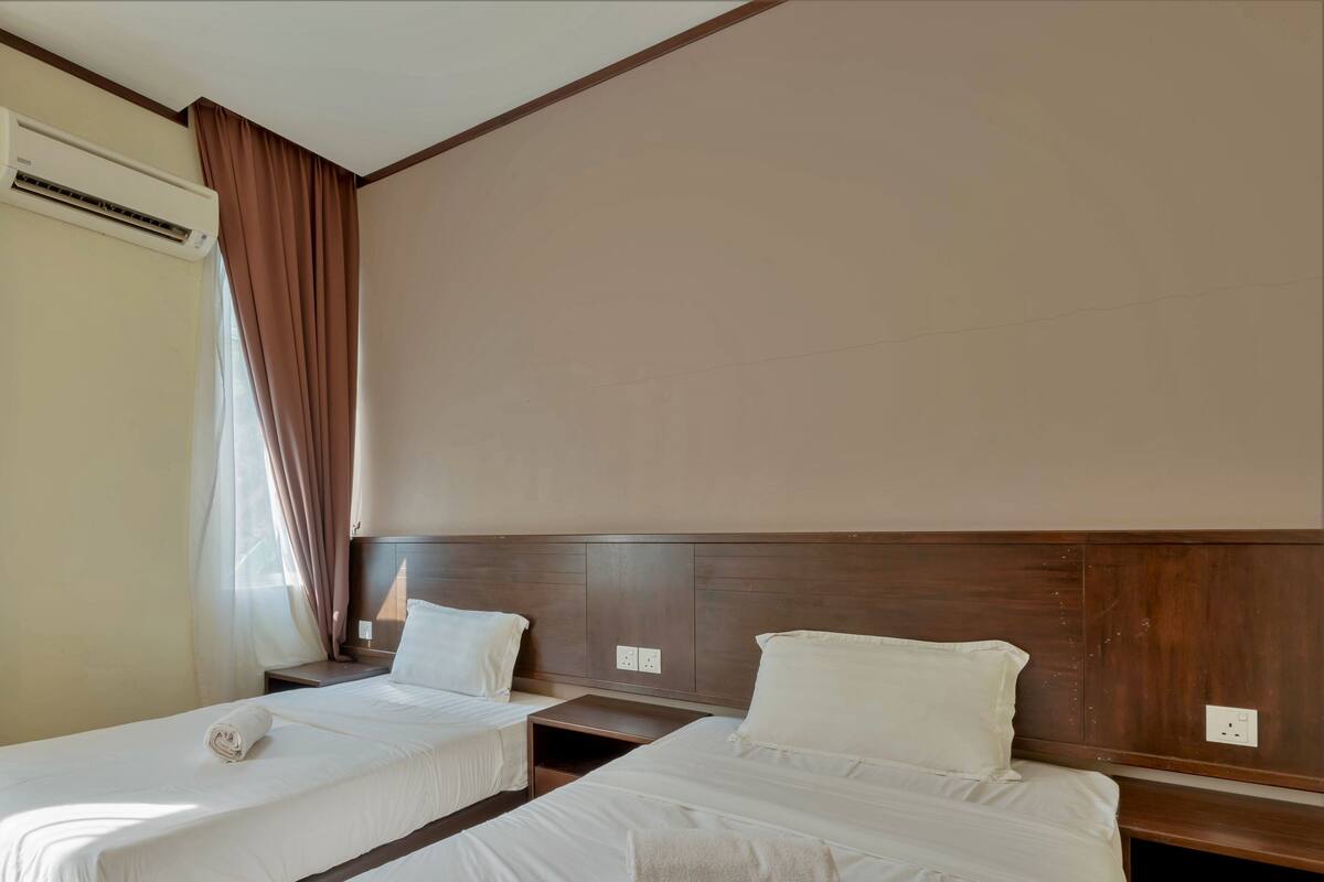A hotel room with two single beds, a wooden headboard, an air conditioning unit, and a window with curtains.