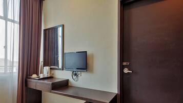 Habitación de hotel con puerta de madera oscura, televisor de pantalla plana montado, un escritorio pequeño con una taza y un teléfono, y una ventana con cortinas translúcidas.