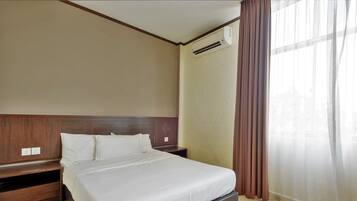 Quarto de hotel com cama, cabeceira de madeira, mesas de cabeceira, um criado-mudo, unidade de ar condicionado e uma janela com cortinas.