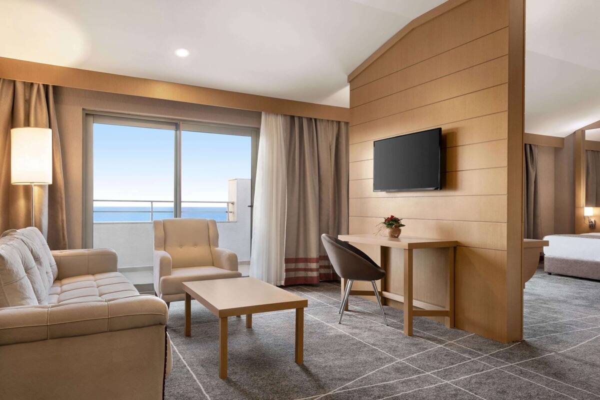 Habitación de hotel con sofá, sillón, escritorio y televisor montado en la pared.