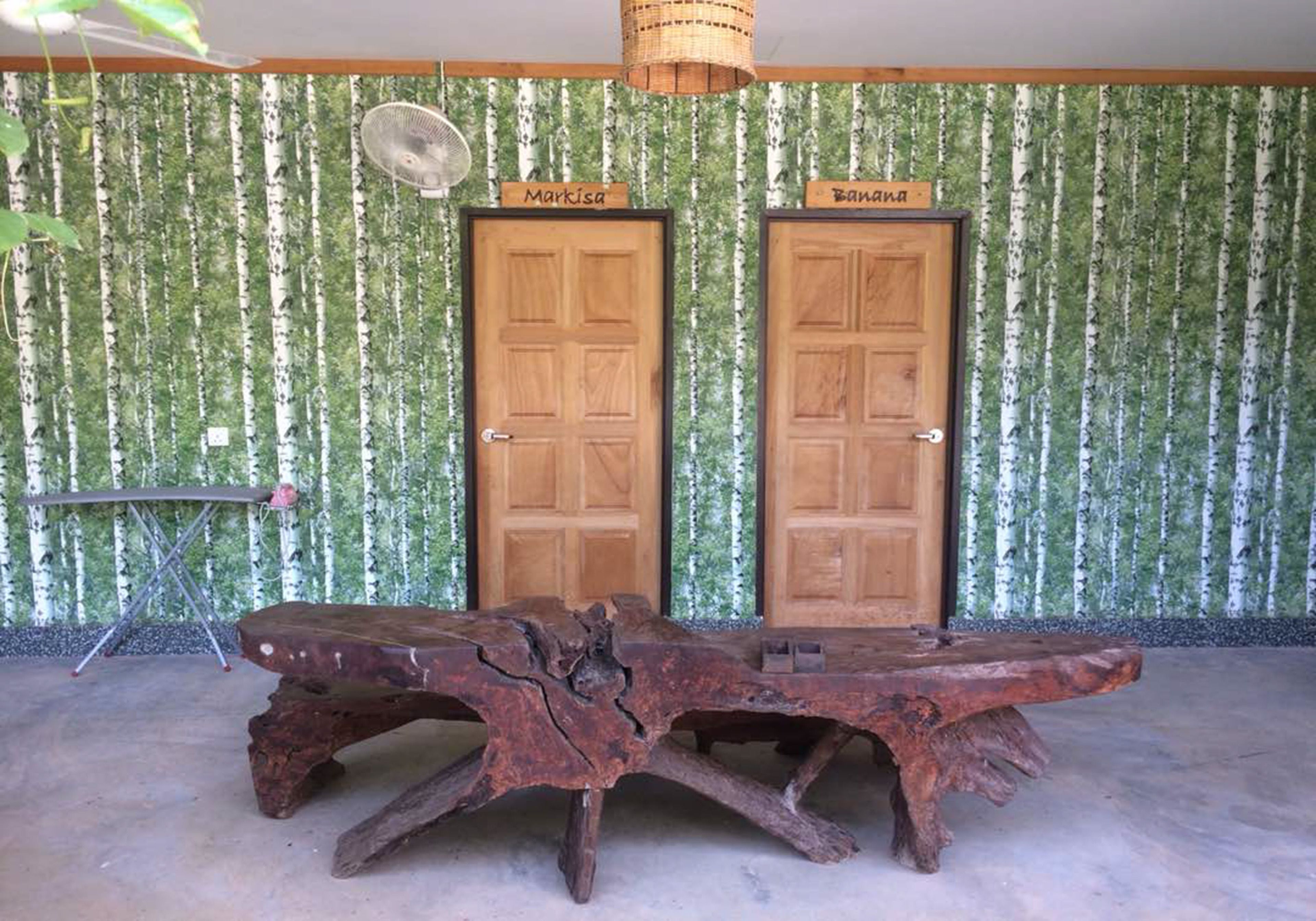 A rustic wooden table with a unique design, two wooden doors labeled 'Markisa' and 'Banana', and a fan against a wall with a birch tree pattern.