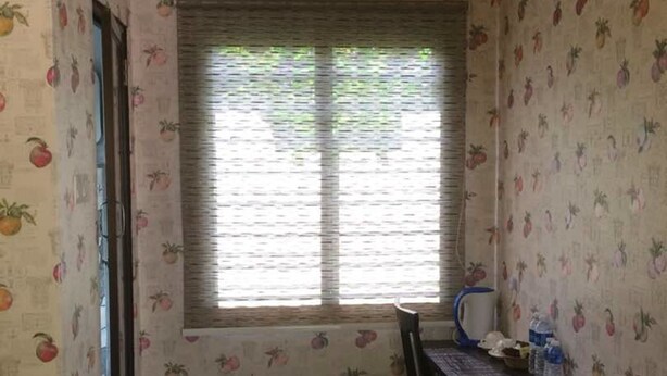 Une petite pièce avec un papier peint à motifs, une fenêtre avec des stores, une chaise, une table et une poubelle.