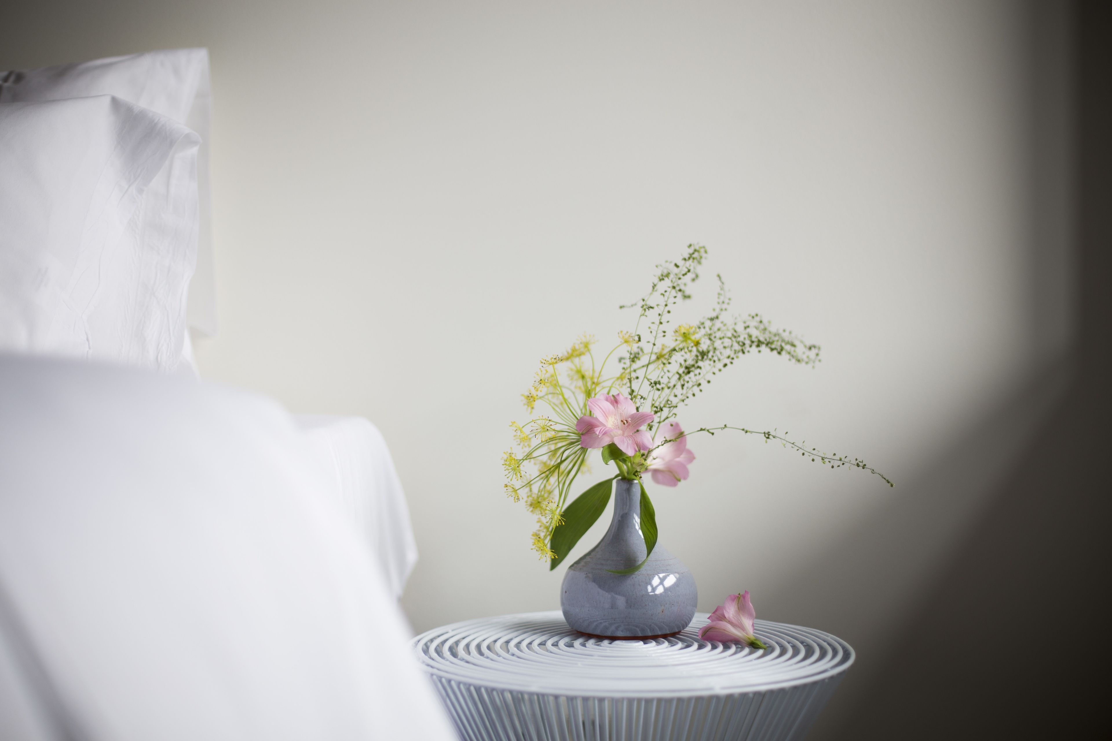 Ein weißes Bett mit einem Kissen, eine graue Vase mit rosa Blumen und Grünpflanzen auf einem runden weißen Tisch.