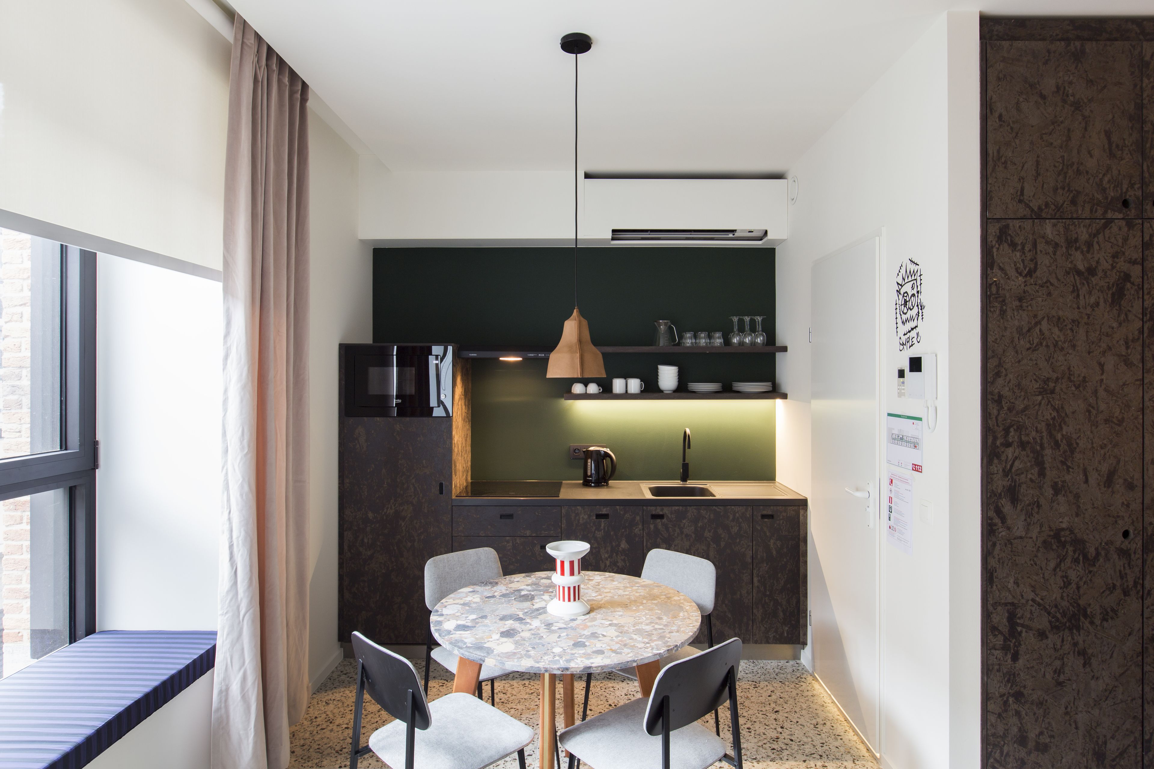A compact kitchen with a dining area, featuring a round table with chairs, a pendant light, and a view of the outside through a window.