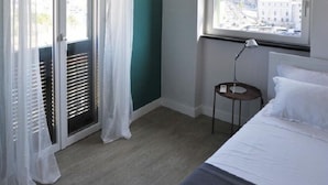 A bedroom with a bed, a window with a view of a cityscape, a nightstand with a lamp, and a balcony with white curtains. - Aquarium Rooms (Genoa)