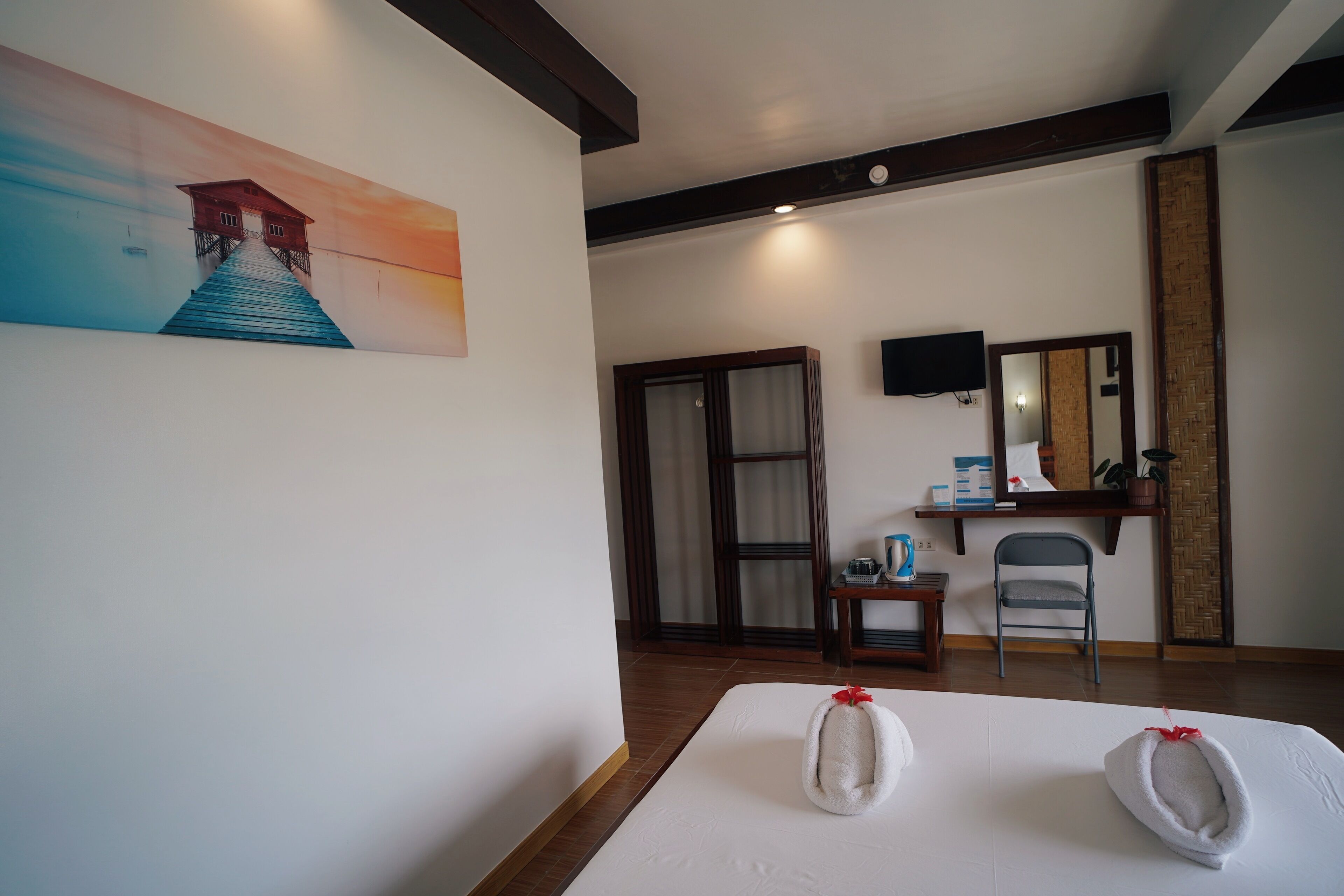 A hotel room with a bed, a painting of a house on a pier, a wooden floor, a chair, a small table with a bottle, and a mirror on the wall.