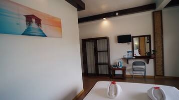 A hotel room with a bed, a painting of a house on a pier, a wooden floor, a chair, a small table with a bottle, and a mirror on the wall.