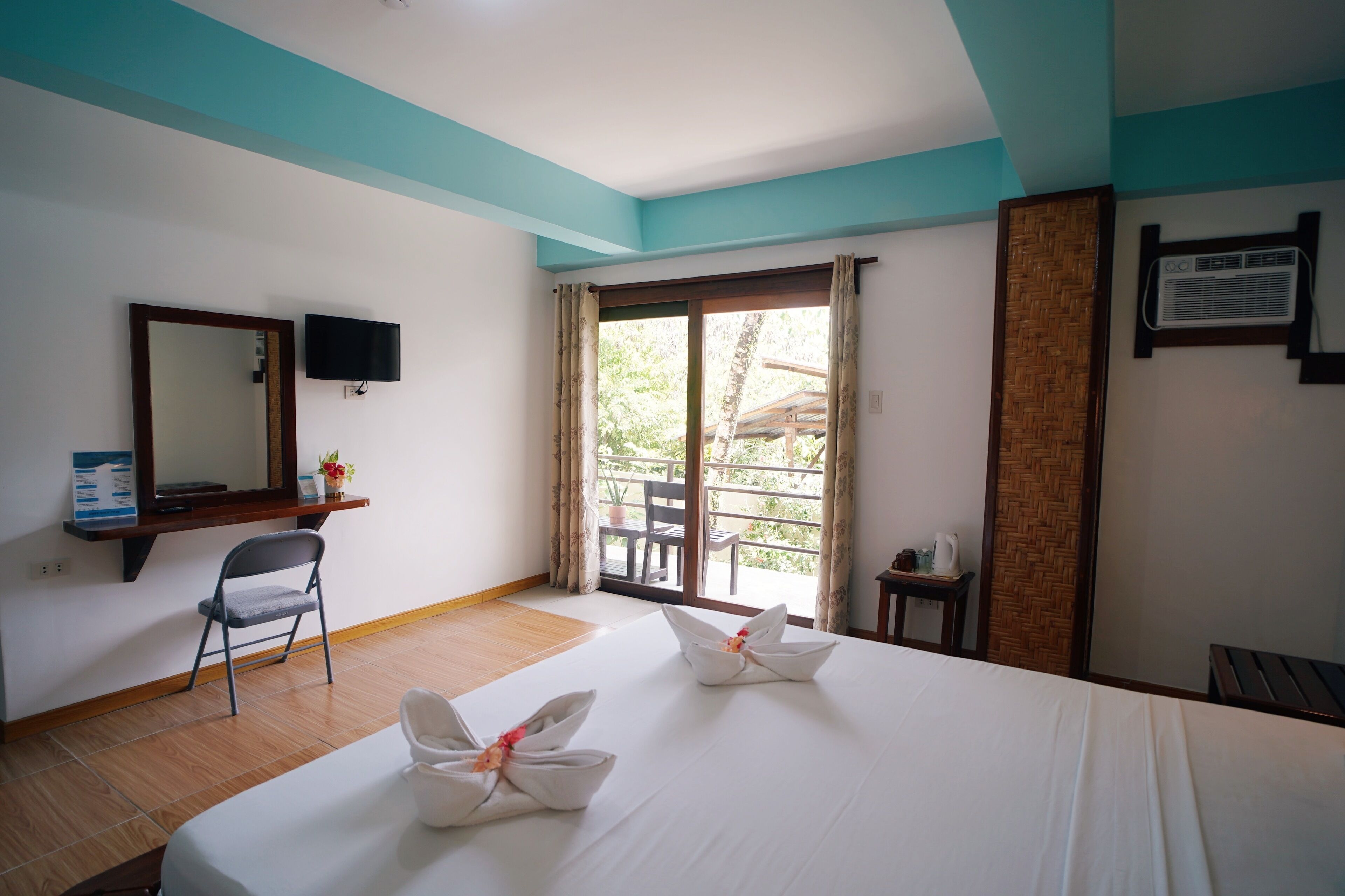 Habitación de hotel con una cama, una silla, televisión, ventana con cortinas y una mesita con hervidor.